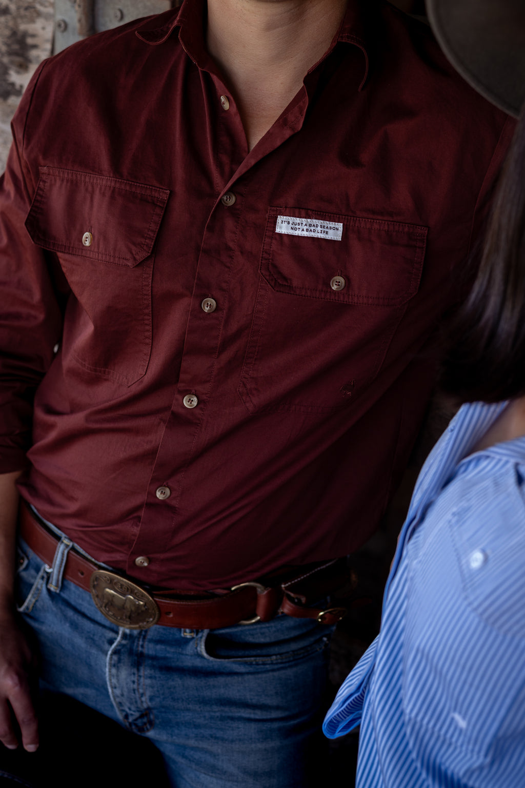 A close-up of a person wearing a burgundy button-down shirt with two chest pockets, paired with blue jeans and a brown belt. A small motivational tag is visible on the left chest pocket.