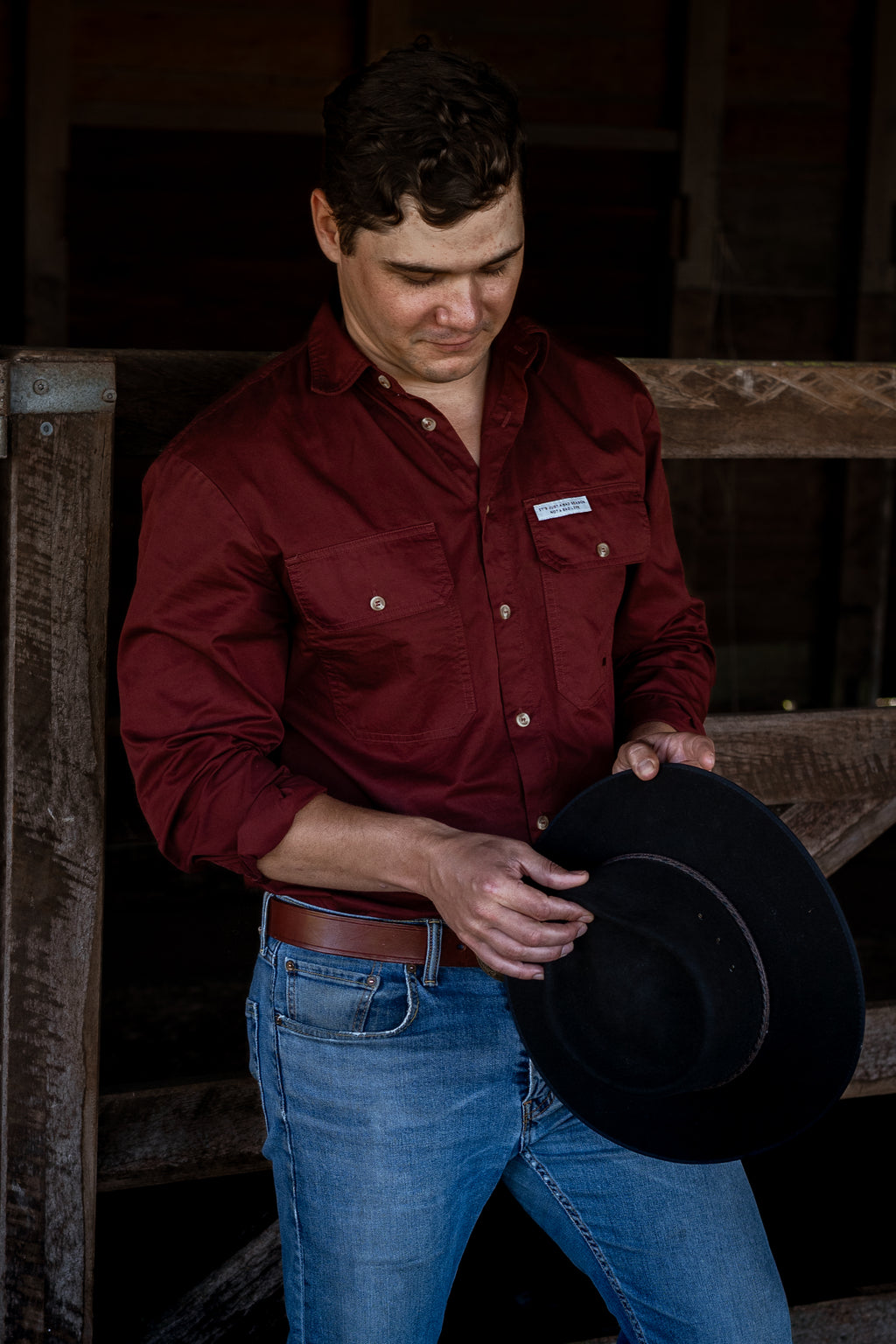 A man wearing a burgundy button-down shirt and blue jeans stands in a rustic barn setting, holding a black cowboy hat.