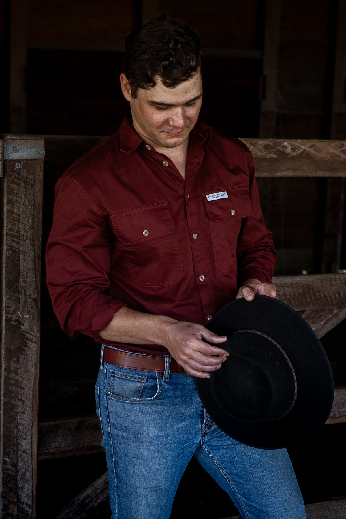 A man wearing a burgundy button-down shirt and blue jeans stands in a rustic barn setting, holding a black cowboy hat.