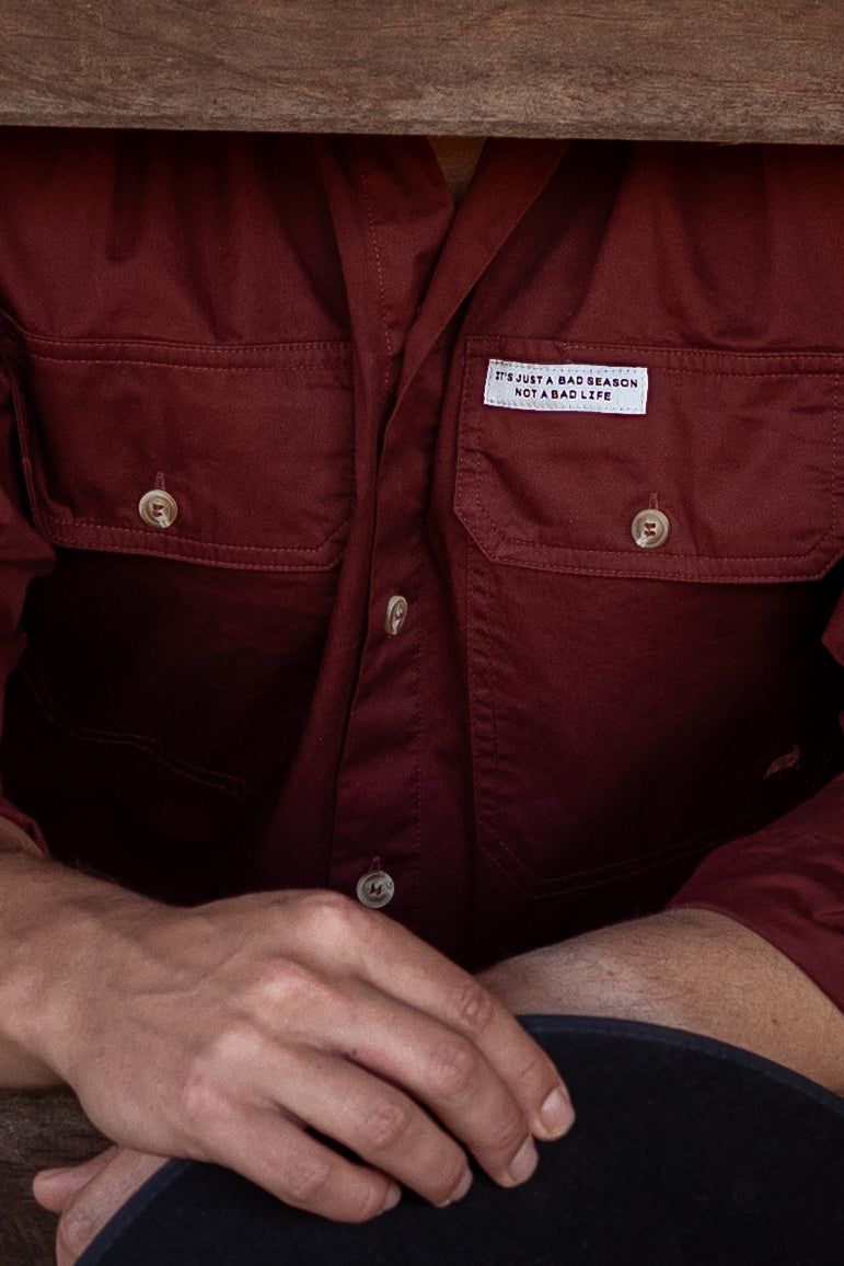 Close-up of a person wearing a dark red button-up shirt with two chest pockets and a white motivational tag. 