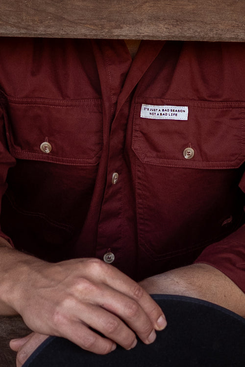 Close-up of a person wearing a dark red button-up shirt with two chest pockets and a white motivational tag. 