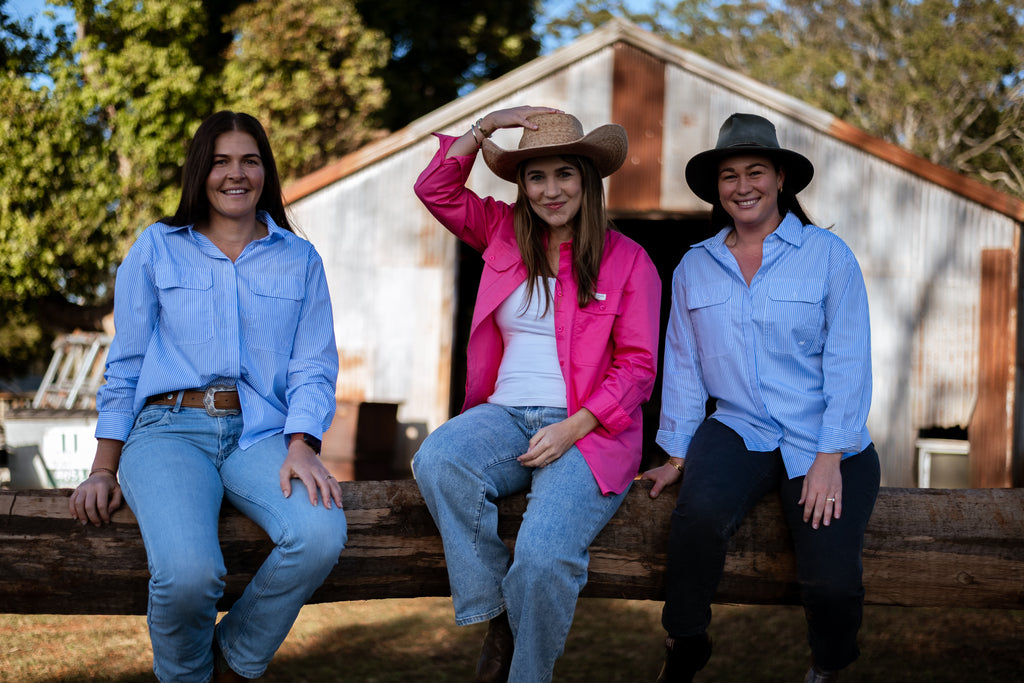 Three women sitting on a wooden fence in front of a rustic corrugated metal barn. The woman on the left wears a light blue button-up shirt and jeans.