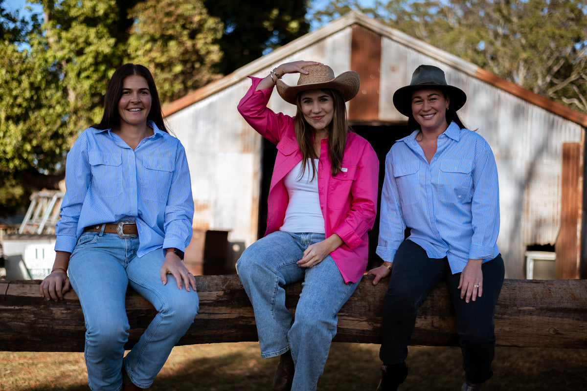 Three women sitting on a wooden fence in front of a rustic corrugated metal barn. The woman on the left wears a light blue button-up shirt and jeans.