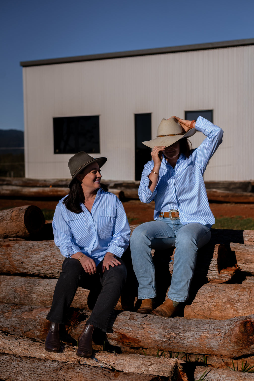 Two women sitting on logs outdoors in front of a white building, wearing light blue button-up shirts and wide-brim hats.
