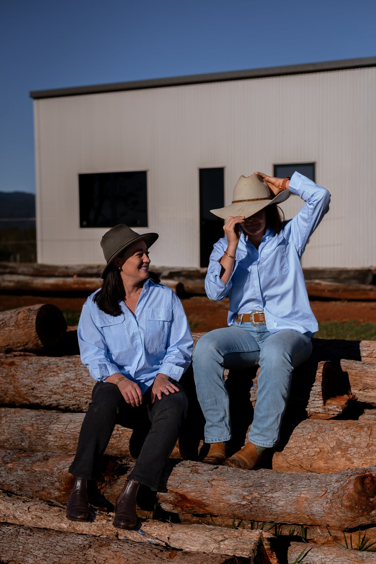 Two women sitting on logs outdoors in front of a white building, wearing light blue button-up shirts and wide-brim hats.