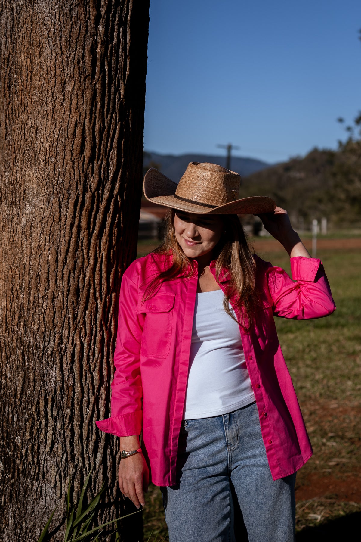 A woman wearing a bright pink button-up shirt, a straw cowboy hat, and a white tank top stands outdoors next to a large tree trunk, with a grassy field and blue sky in the background.