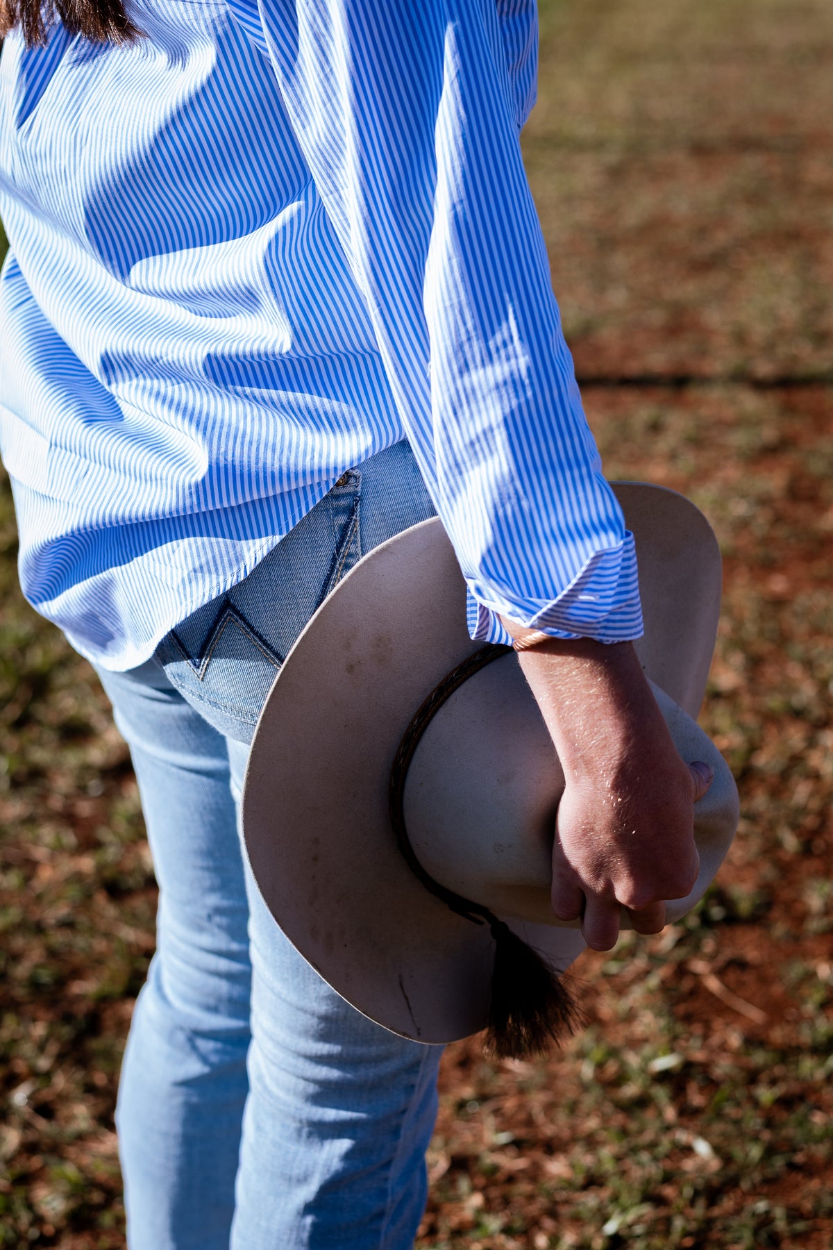 A person outdoors wearing a light blue and white striped long-sleeve shirt and light blue jeans, holding a beige cowboy hat in one hand.