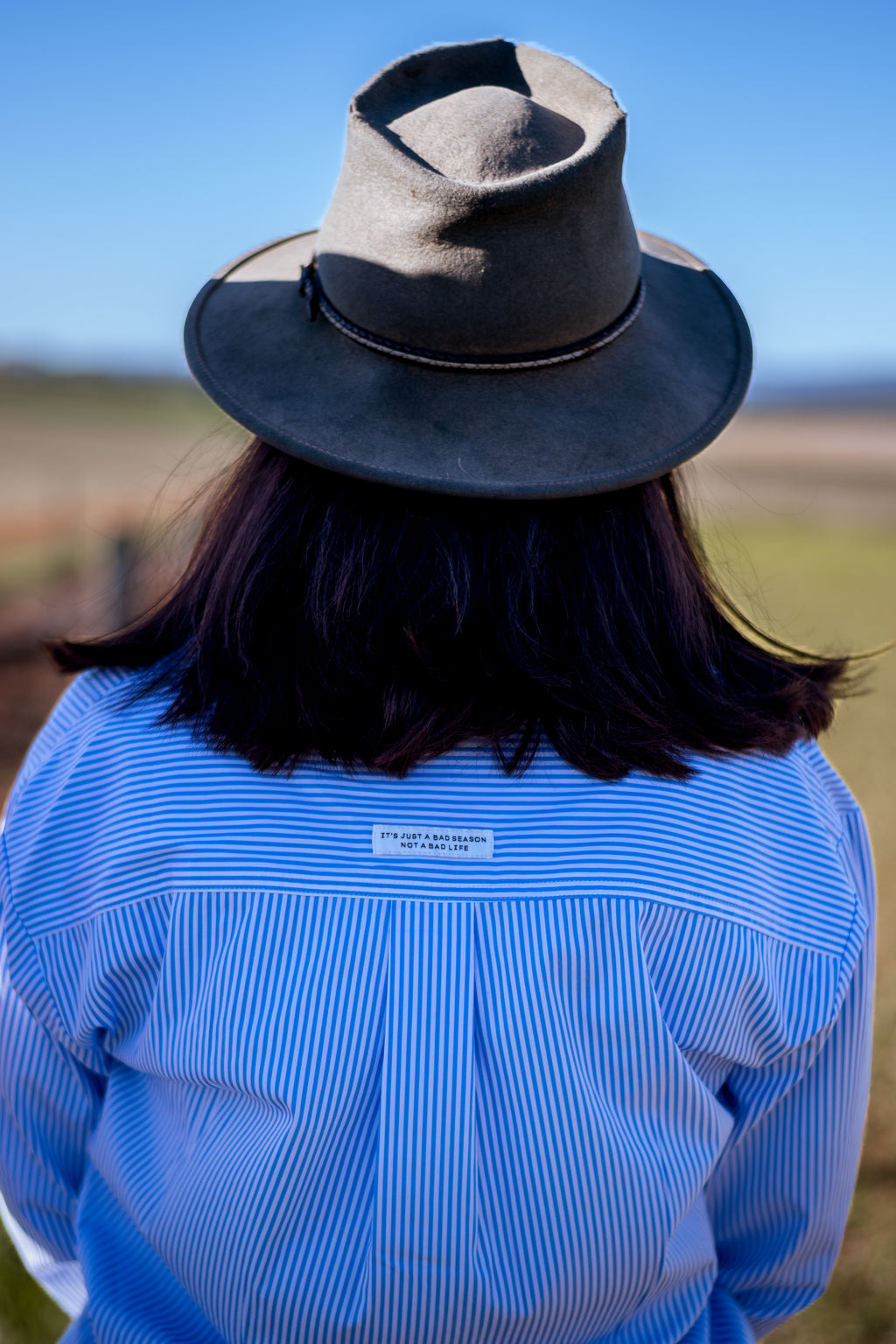A person viewed from behind wearing a dark gray felt wide-brim hat and a light blue and white vertically striped button-down shirt, standing outdoors against a blue sky and blurred grassy field.