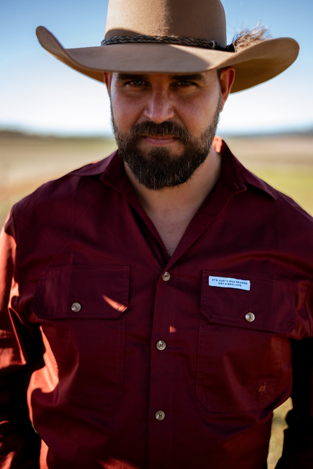 A man wearing a tan cowboy hat and a burgundy button-up shirt with a visible Columbia logo patch, photographed outdoors against a blurred natural background.