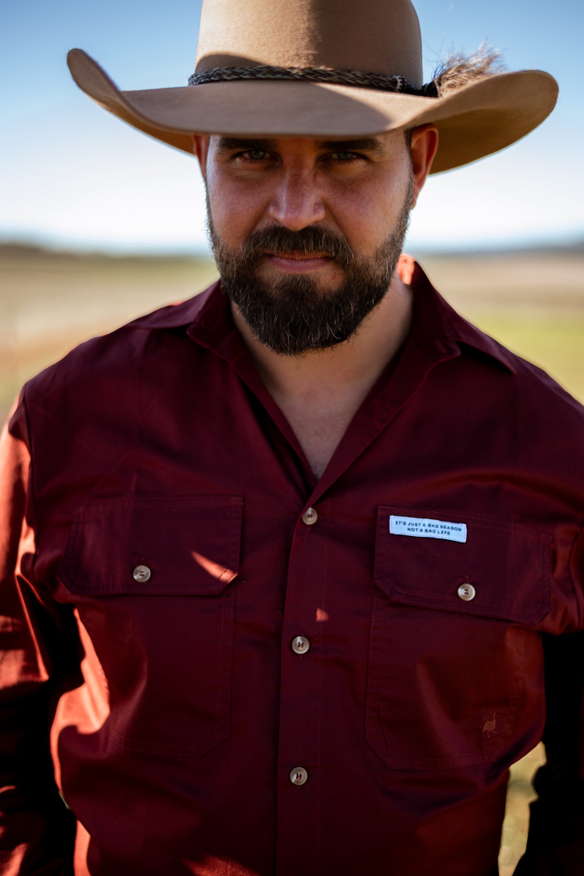 A man wearing a tan cowboy hat and a burgundy button-up shirt with a visible Columbia logo patch, photographed outdoors against a blurred natural background.