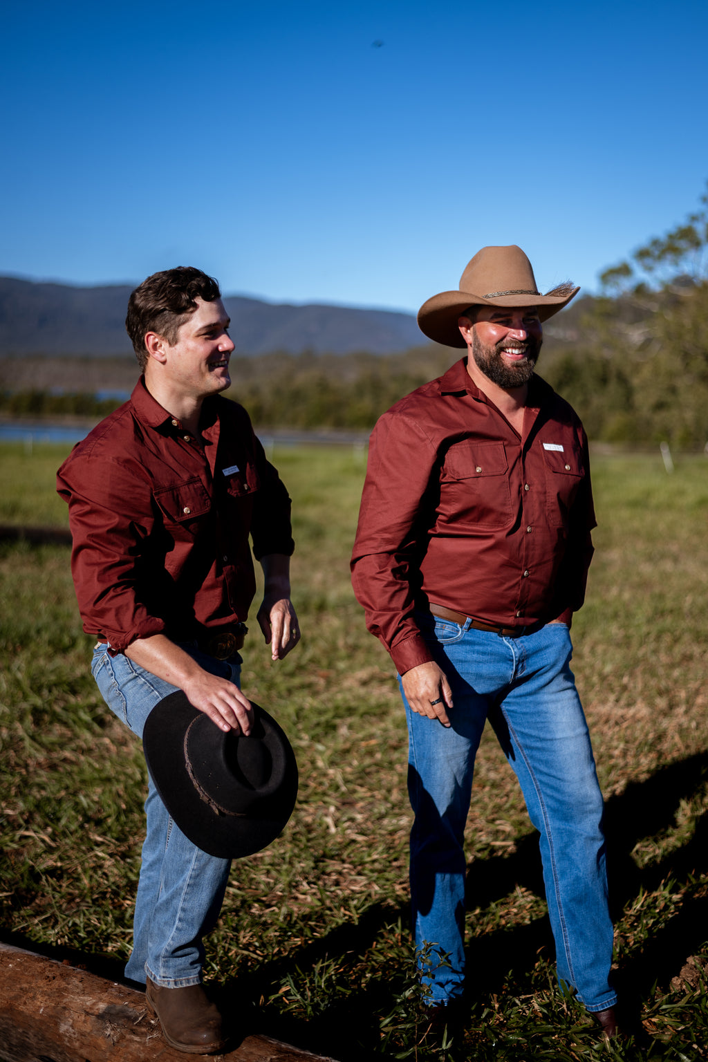 Two men standing outdoors in a grassy field under a blue sky, wearing matching burgundy button-up shirts and jeans. One man holds a black cowboy hat, and the other wears a tan cowboy hat.