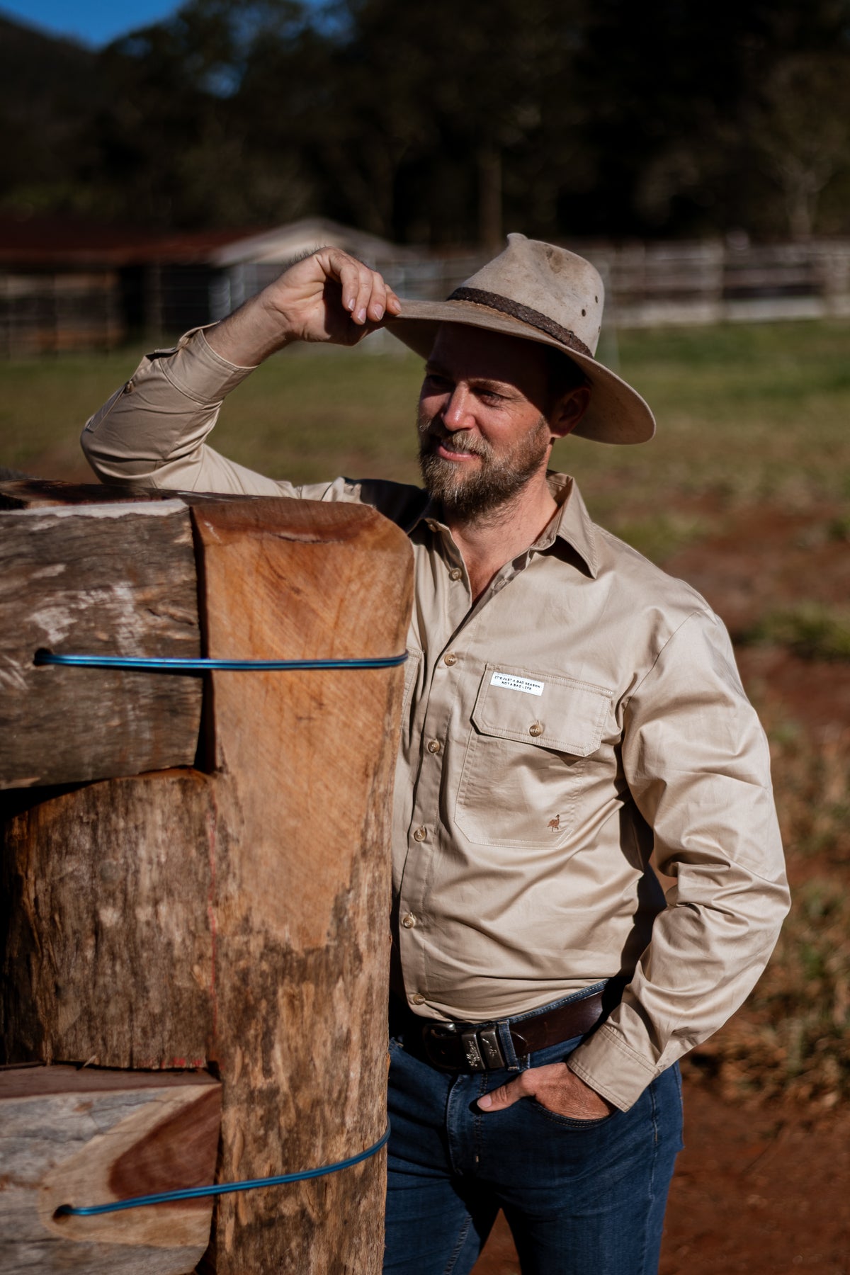 A man wearing a beige long-sleeve button-up shirt and a wide-brimmed hat leans against a rustic wooden post in an outdoor farm setting.