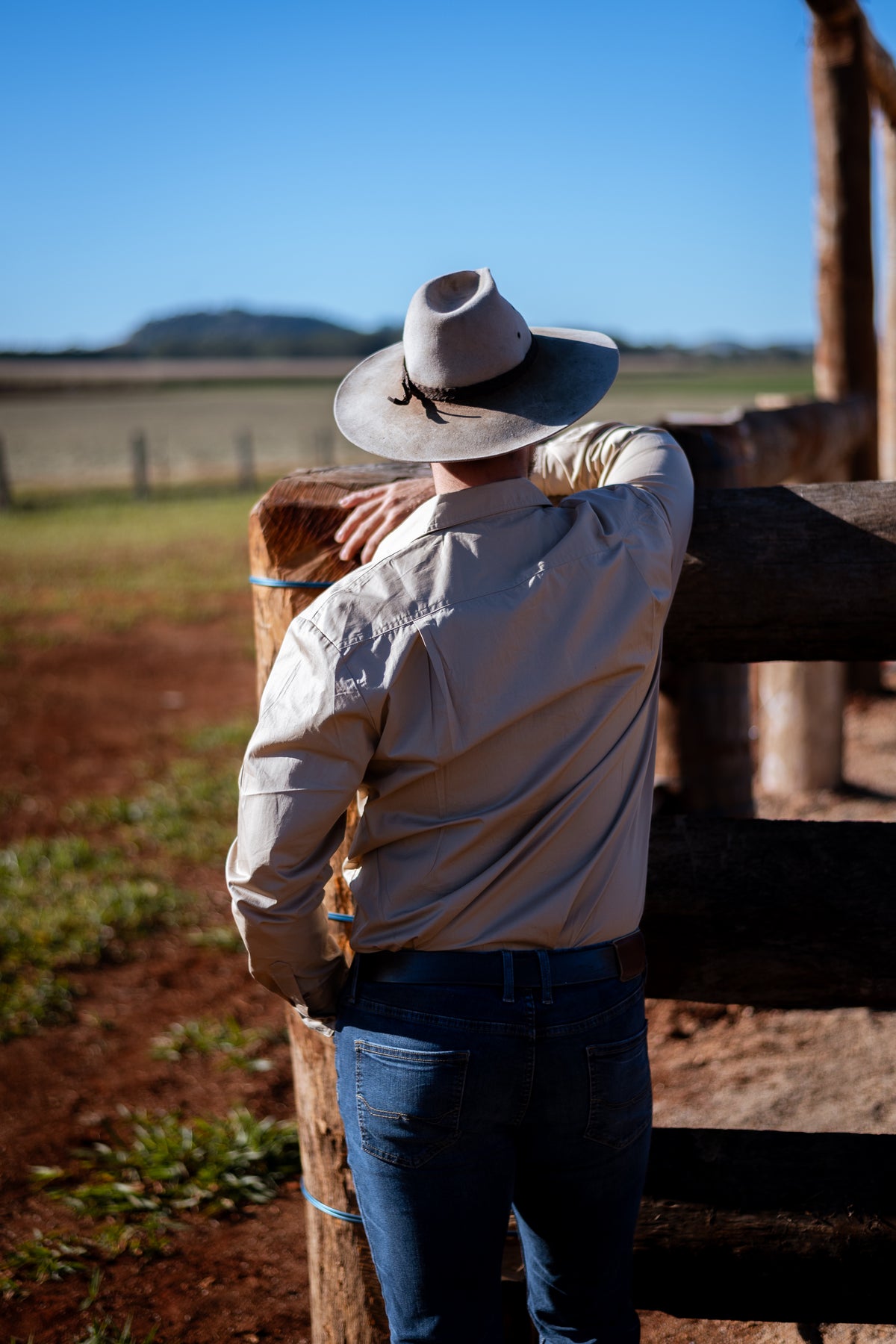 A man wearing a light beige cowboy hat, a beige long-sleeve button-up shirt, and blue jeans stands with his back to the camera, leaning on a wooden fence in an outdoor ranch setting under a clear blue sky.