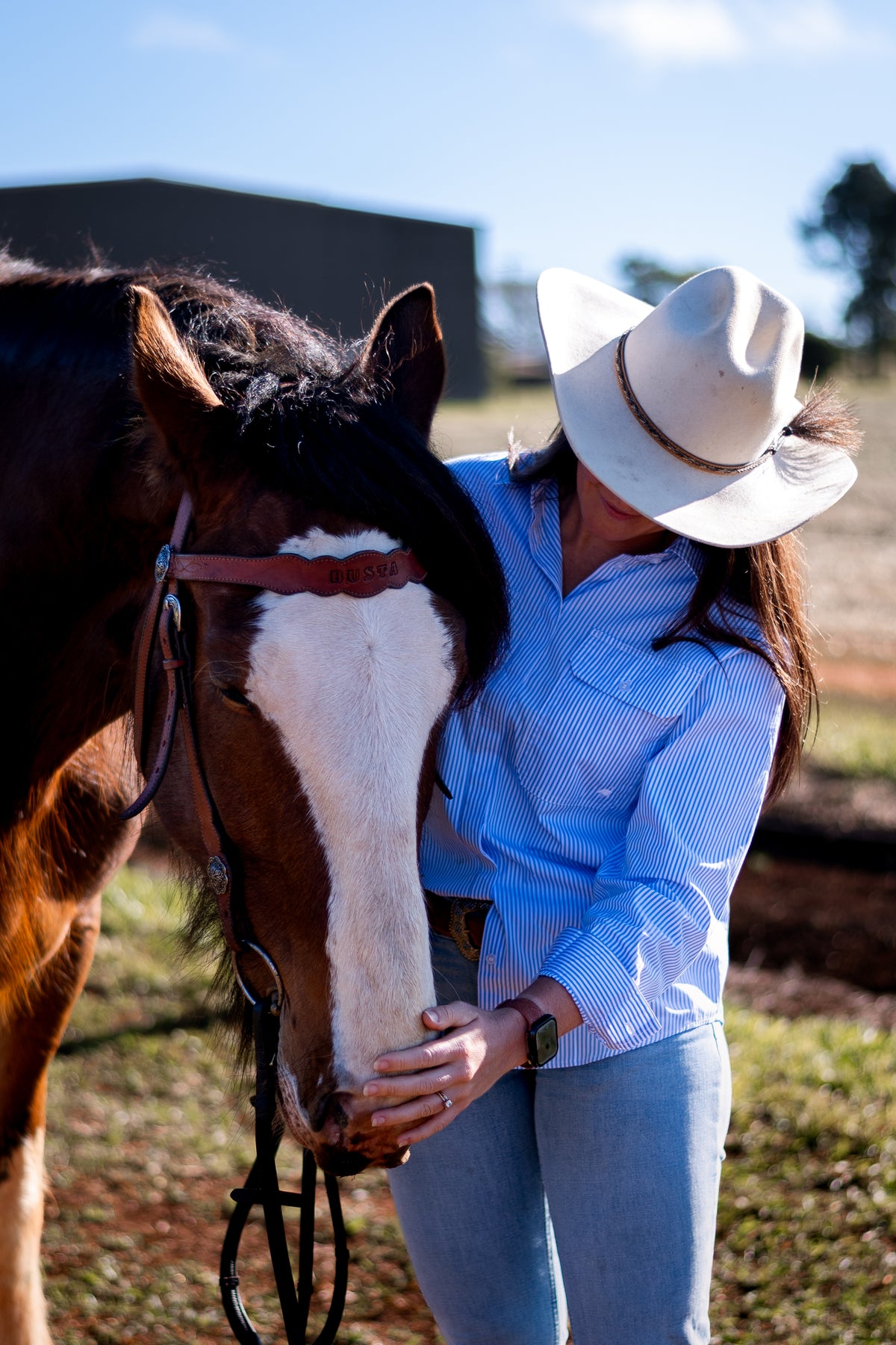 A woman in a white cowboy hat, light blue striped shirt, and jeans stands beside a brown and white horse outdoors, touching its face. The background shows a dirt ground, grass, and a dark building under a blue sky.