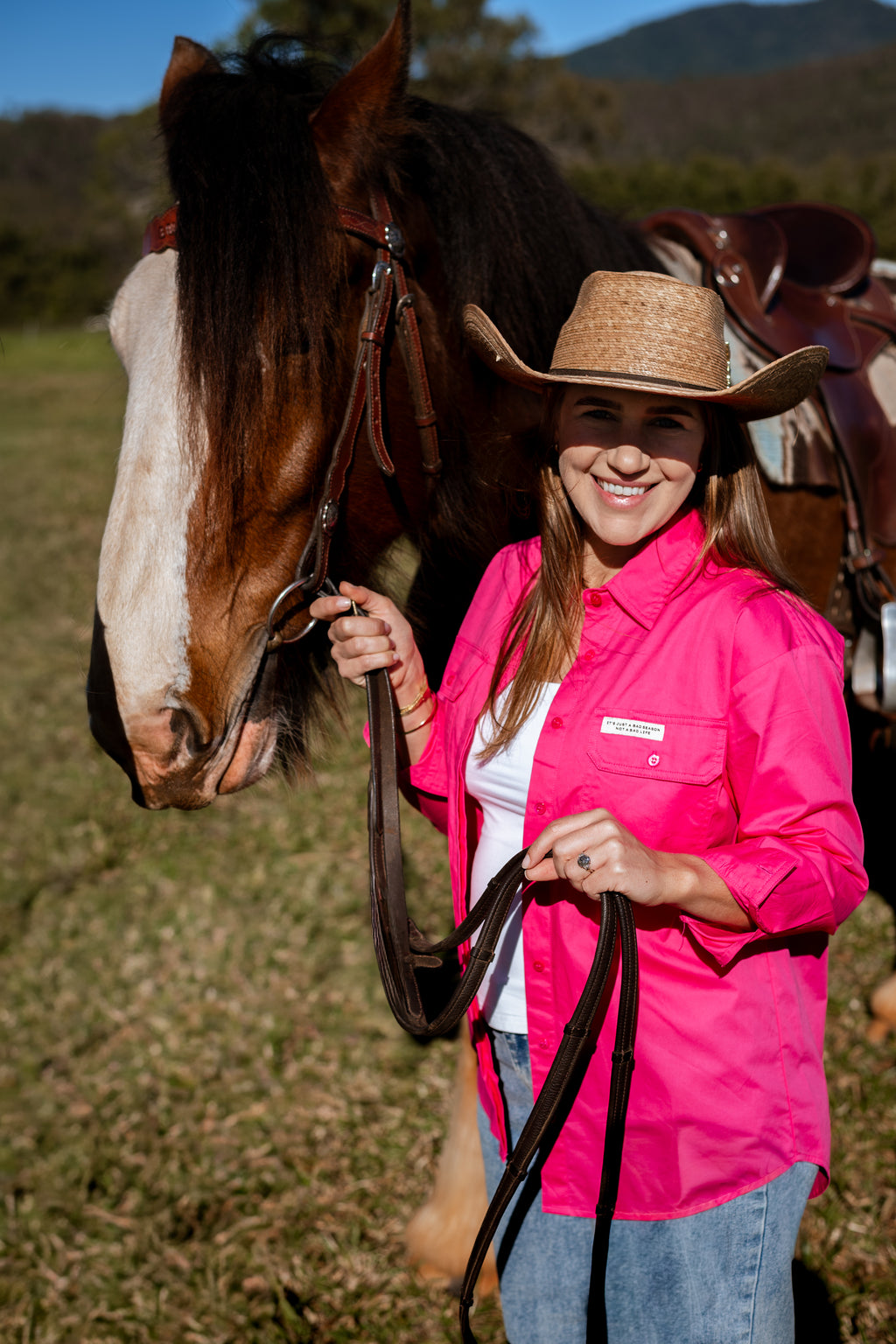 A smiling woman in a cowboy hat and bright pink shirt stands outdoors beside a brown horse, holding its reins in a grassy field.