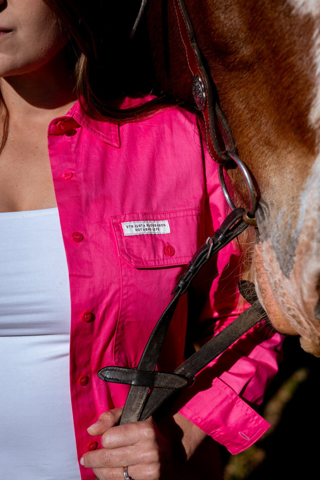 A close-up lifestyle shot of a woman wearing a vibrant hot pink button-up shirt with a chest pocket, holding horse reins next to a brown horse.