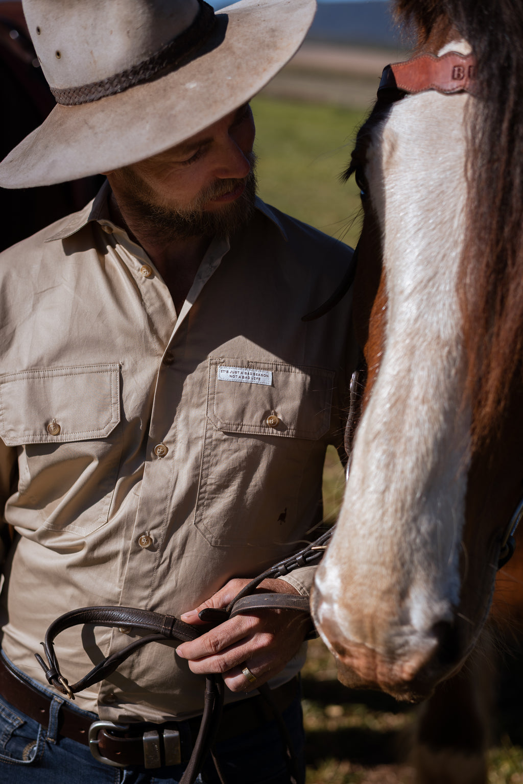 A man in a beige western shirt and cowboy hat standing beside a horse outdoors, holding horse reins. The horse has a white and brown coat and a leather bridle.