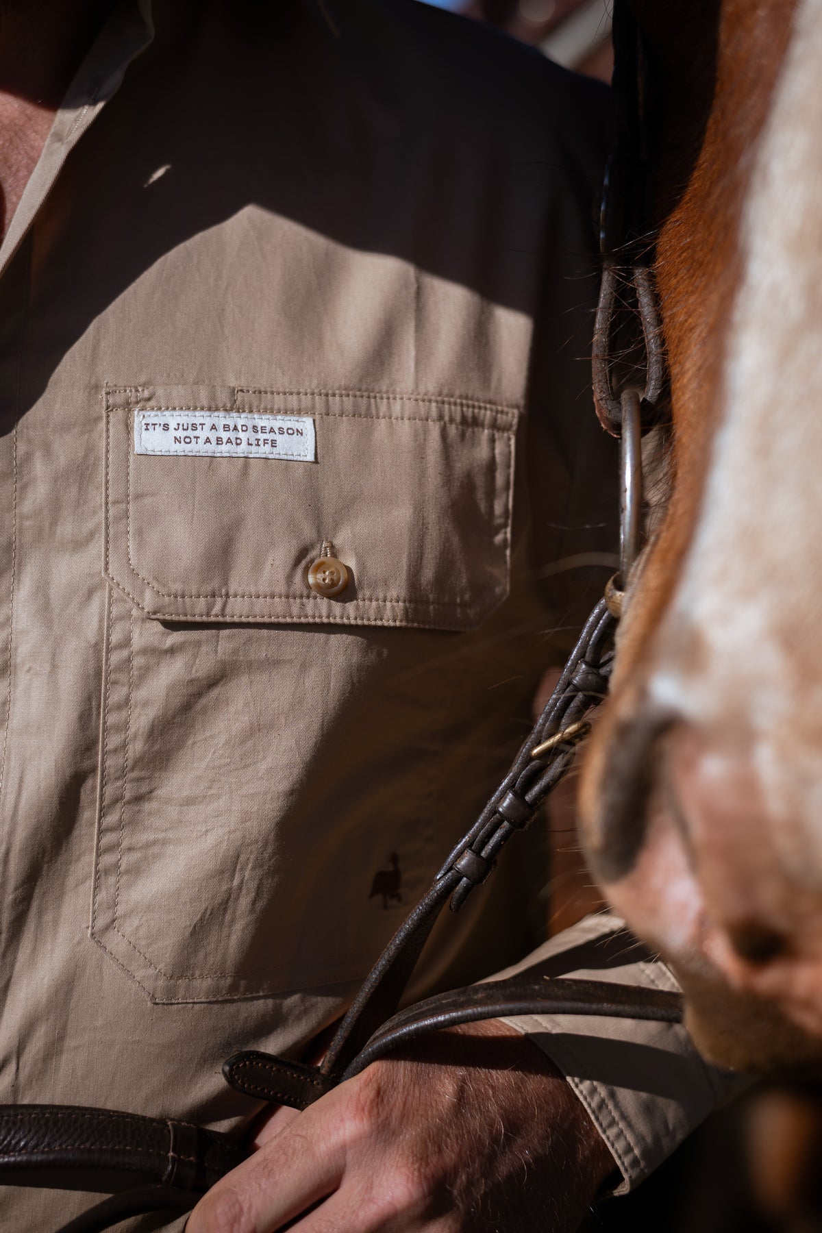 Close-up of a person wearing a tan buttoned chest pocket shirt with a visible motivational tag.